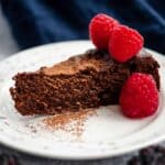slice of flourless chocolate cake with a sprinkle of cocao powder and topped with 3 raspberries