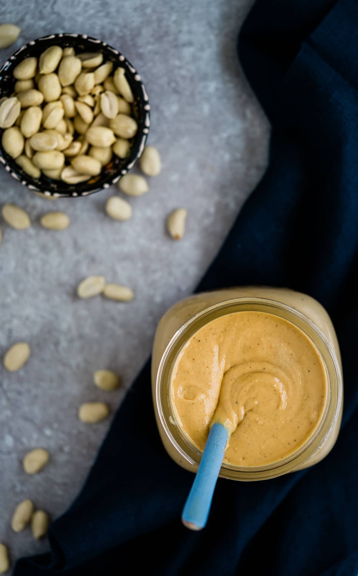 Homemade peanut butter in a jar with a blue spoon and a side of roasted peanuts in a small bowl