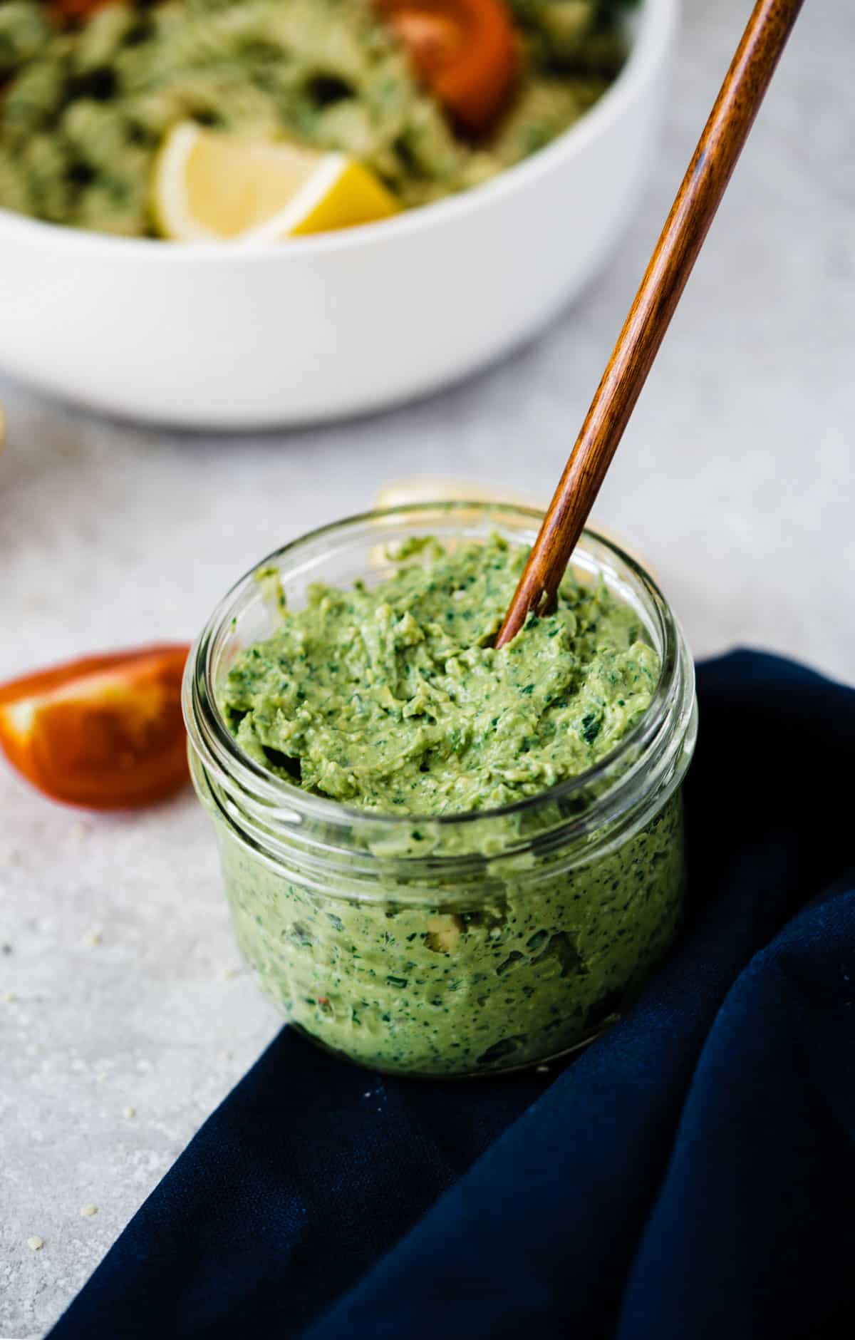 Vegan avocado and basil pesto in a mason jar with a spoon and a bowl of pesto pasta in the background
