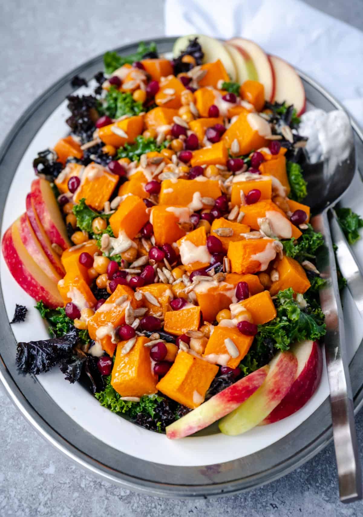 roasted butternut squash, kale and chickpea salad with apple slices, pomegranate and sunflower seeds on a white serve dish