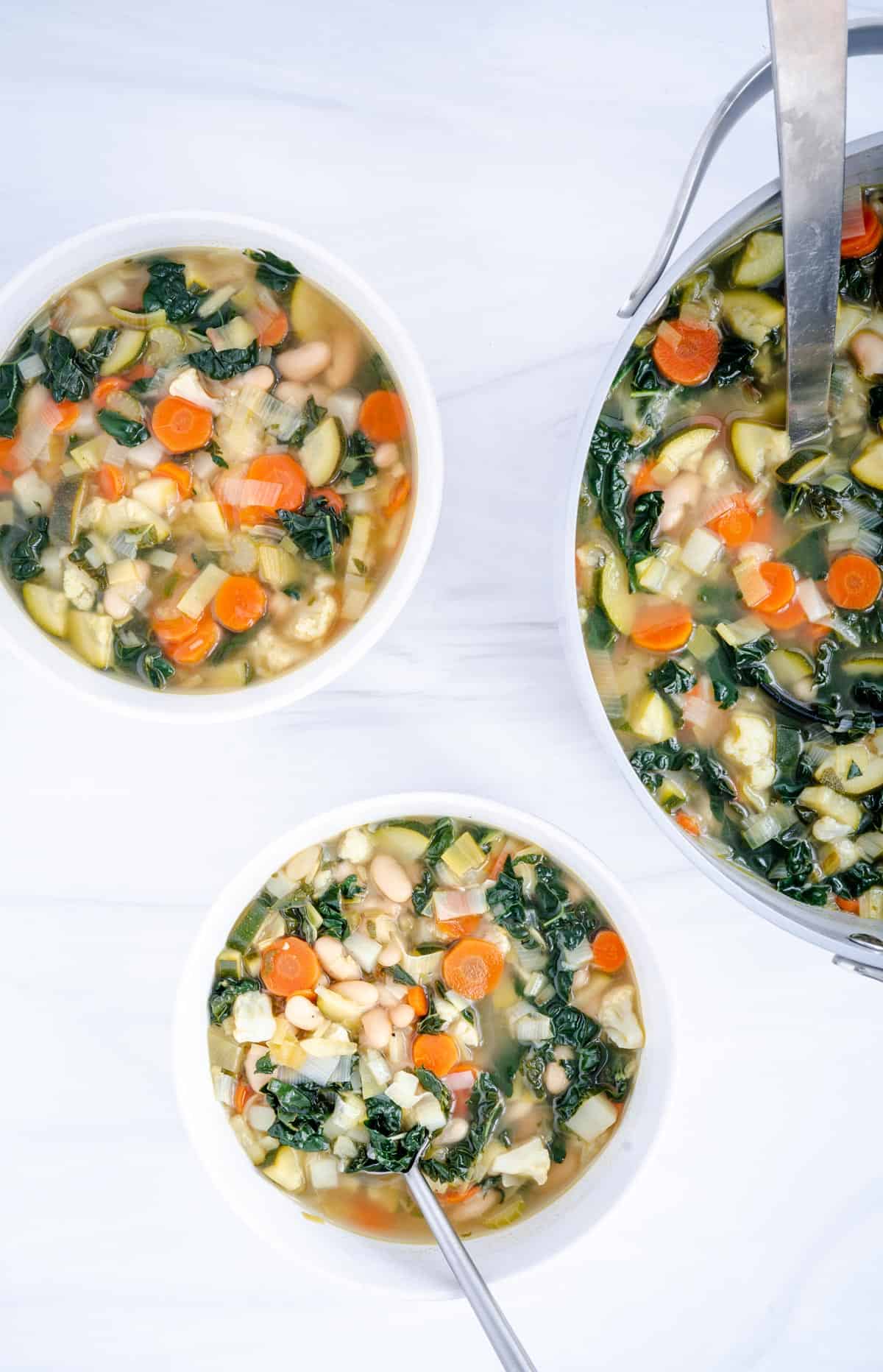 white bean vegetable soup with kale in two white bowl one with spoon and a grey pot with ladle 