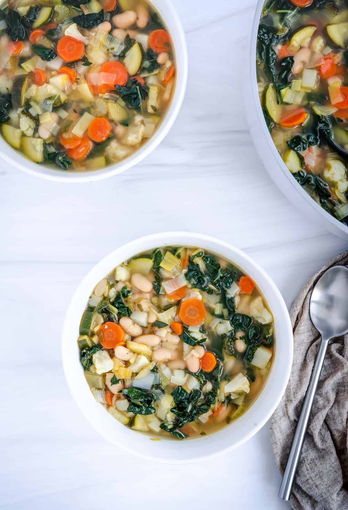 white bean and vegetable soup with kale in two white bowls and a grey pot with a spoon on the side