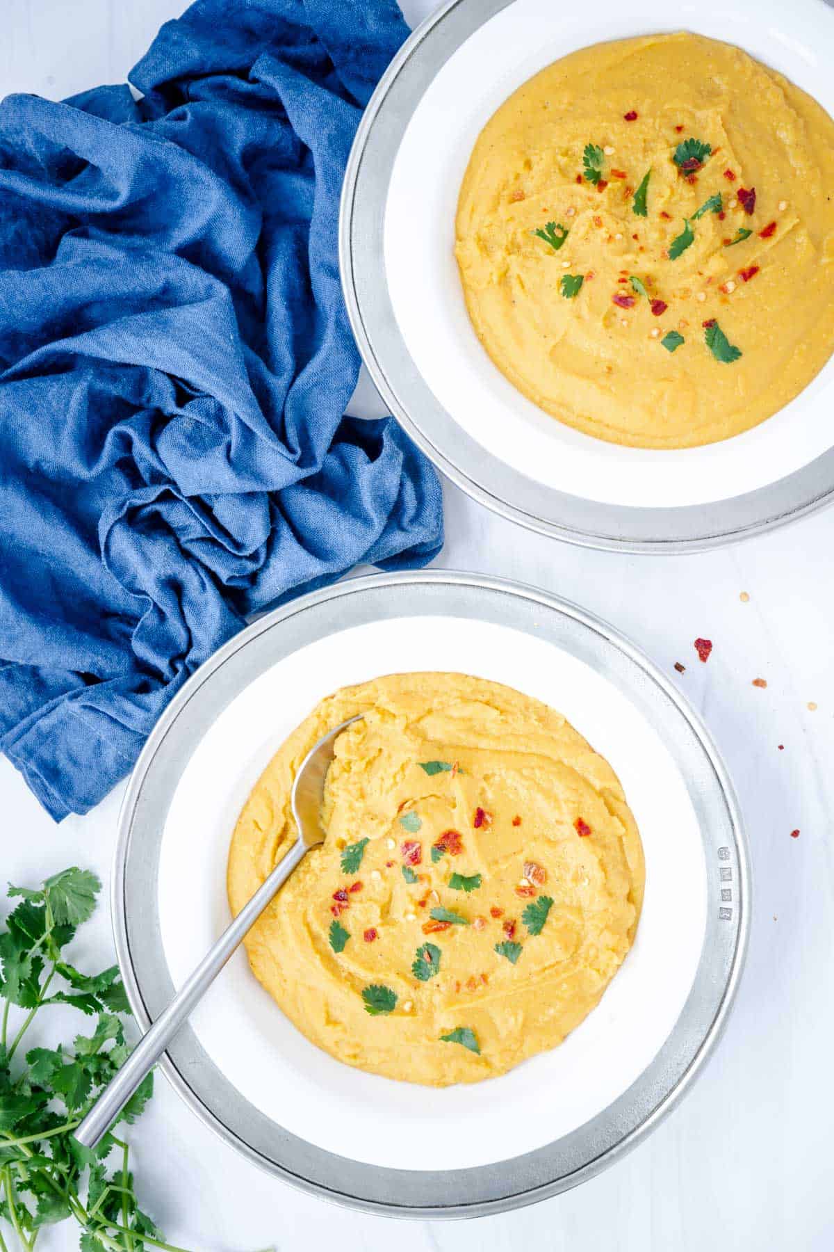 creamy yellow split pea soup with turmeric in two pewter rimmed bowls with spoon blue cloth and cilantro
