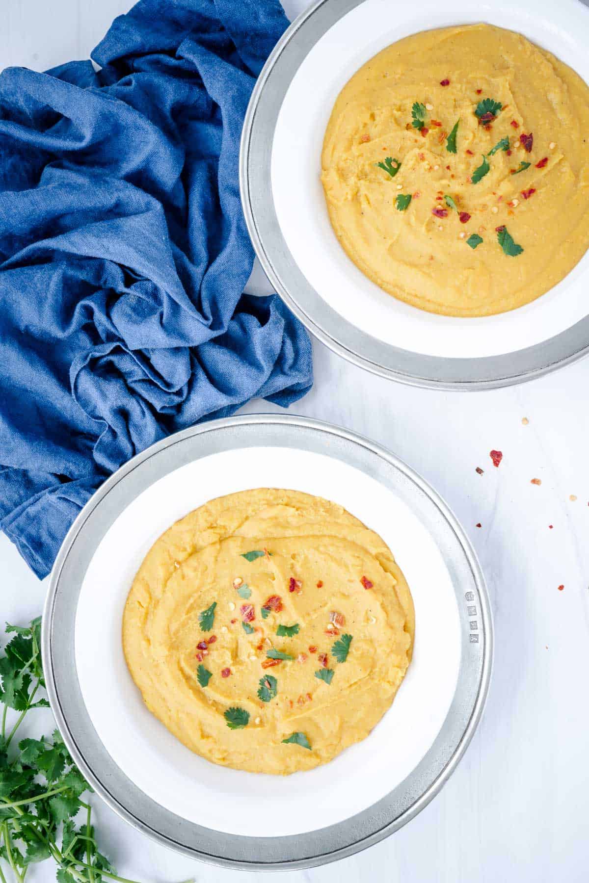 creamy yellow split pea soup with turmeric in two bowls sprinkled with red pepper flakes and cilantro beside a blue cloth