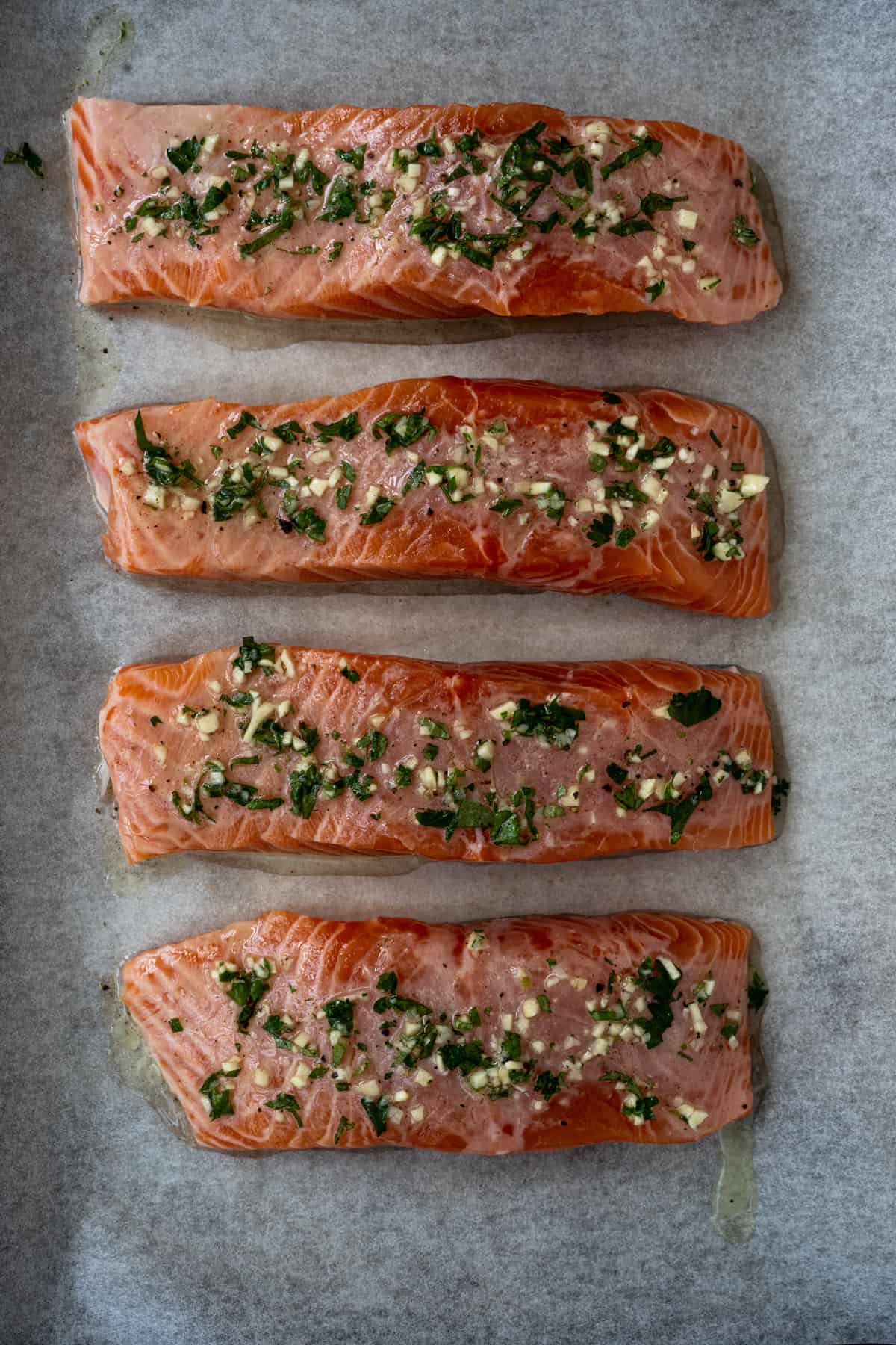 Four filets of raw salmon topped with lemon garlic herb on parchment lined bake tray.