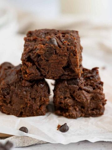 sweet potato brownies piled in pyramid of 3 brownies on parchment paper on wood board with beige napkin in background.