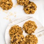 Five banana oatmeal breakfast cookies piled on a white plate with a few cookies around and a scattering of oats.
