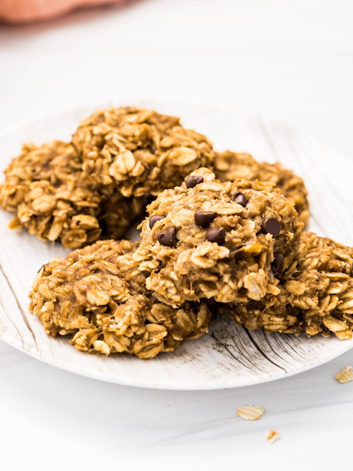 Six banana oatmeal breakfast cookies piled on a white plate with a pink napkin in the background.