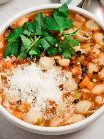 White bowl filled with gluten free pasta fagioli topped with grated parmesan and chopped parsley with a spoon on the side.