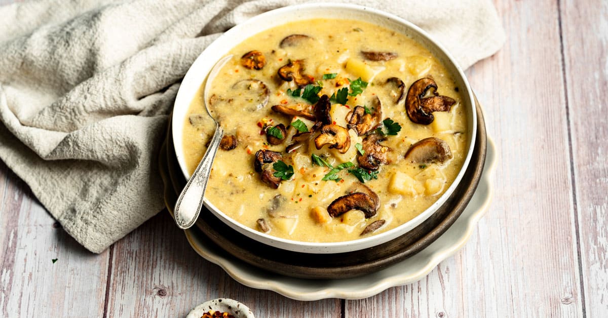 Mushroom potato soup in white bowl with spoon on brown charger with beige napkin.