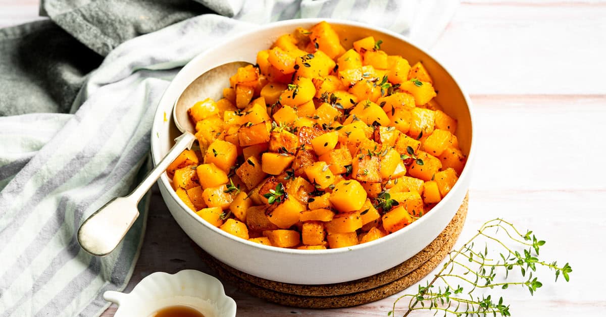 Sauteed butternut squash in a white bowl with serving spoon and striped napkin.