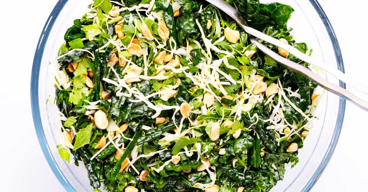 Kale salad with peanut dressing in a clear bowl with silver salad tongs.