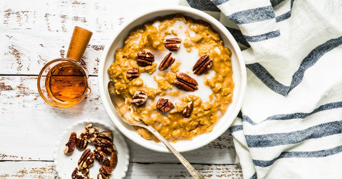 Pumpkin oatmeal in white bowl with spoon with small dish of pecans on the side and blue napkin.