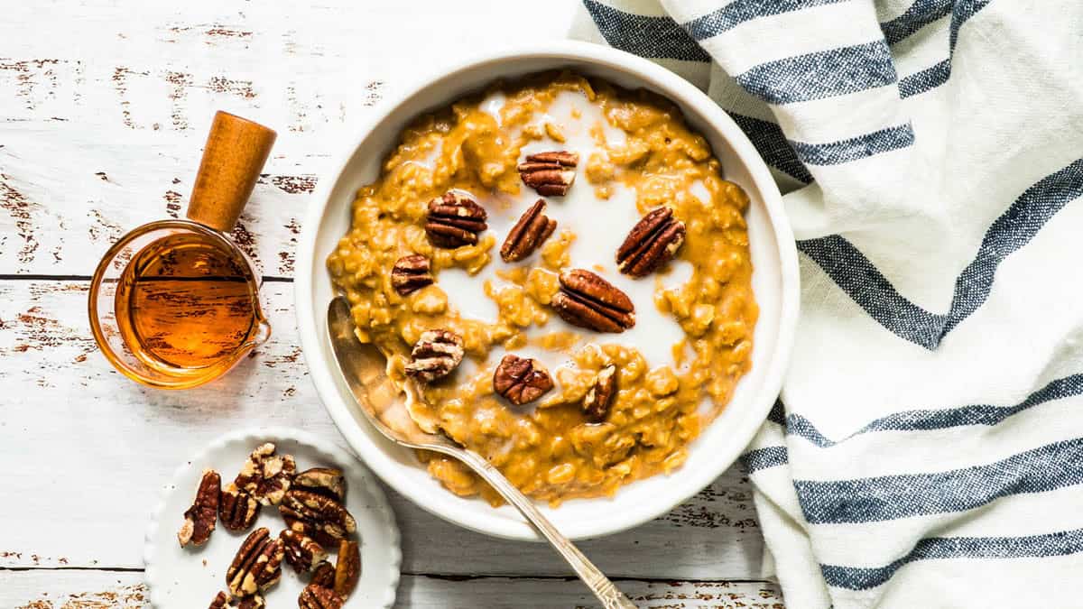 Pumpkin oatmeal with spoon in a white bowl with maple syrup, chopped pecans and a striped napkin on the side.