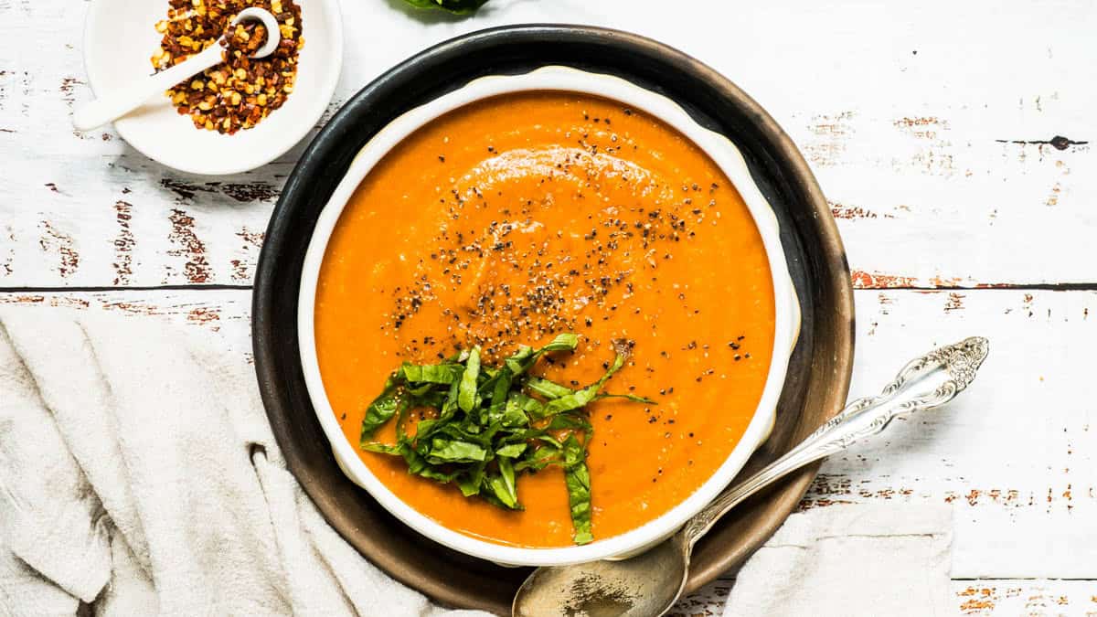 Tomato soup with basil in soup bowl with spoon and beige napkin.